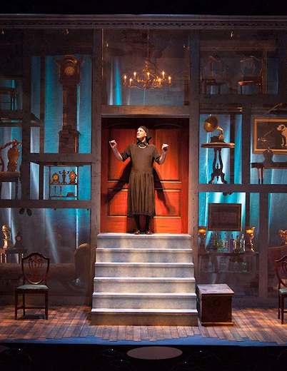 A woman stands confidently on stage, with an open door behind her, suggesting a moment of opportunity or transition.