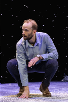 A man kneels on the snowy ground, surrounded by gently falling snowflakes.