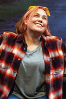 A woman wearing a plaid shirt and jeans sits on a bench, looking thoughtfully into the distance.